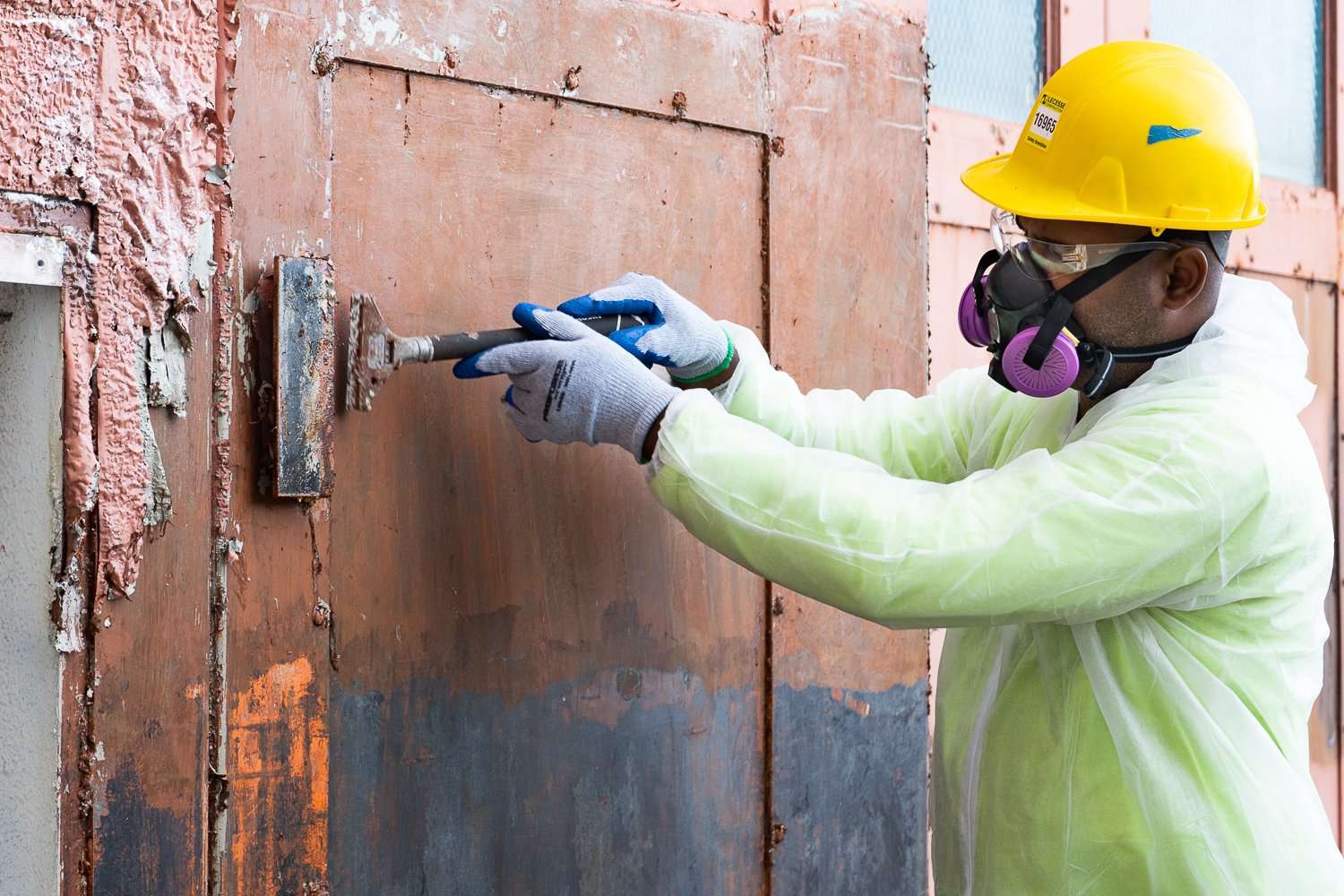 Worker in PPE, hard hat and respirator scraping lead-based paint from a door during compliant removal work