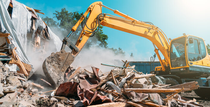 Yellow excavator demolishing a building on a construction site, generating dust and debris